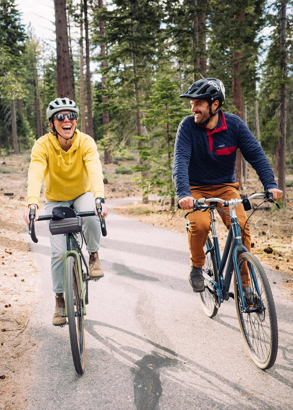Two smiling people riding bikes on a paved path, surrounded by pine trees.