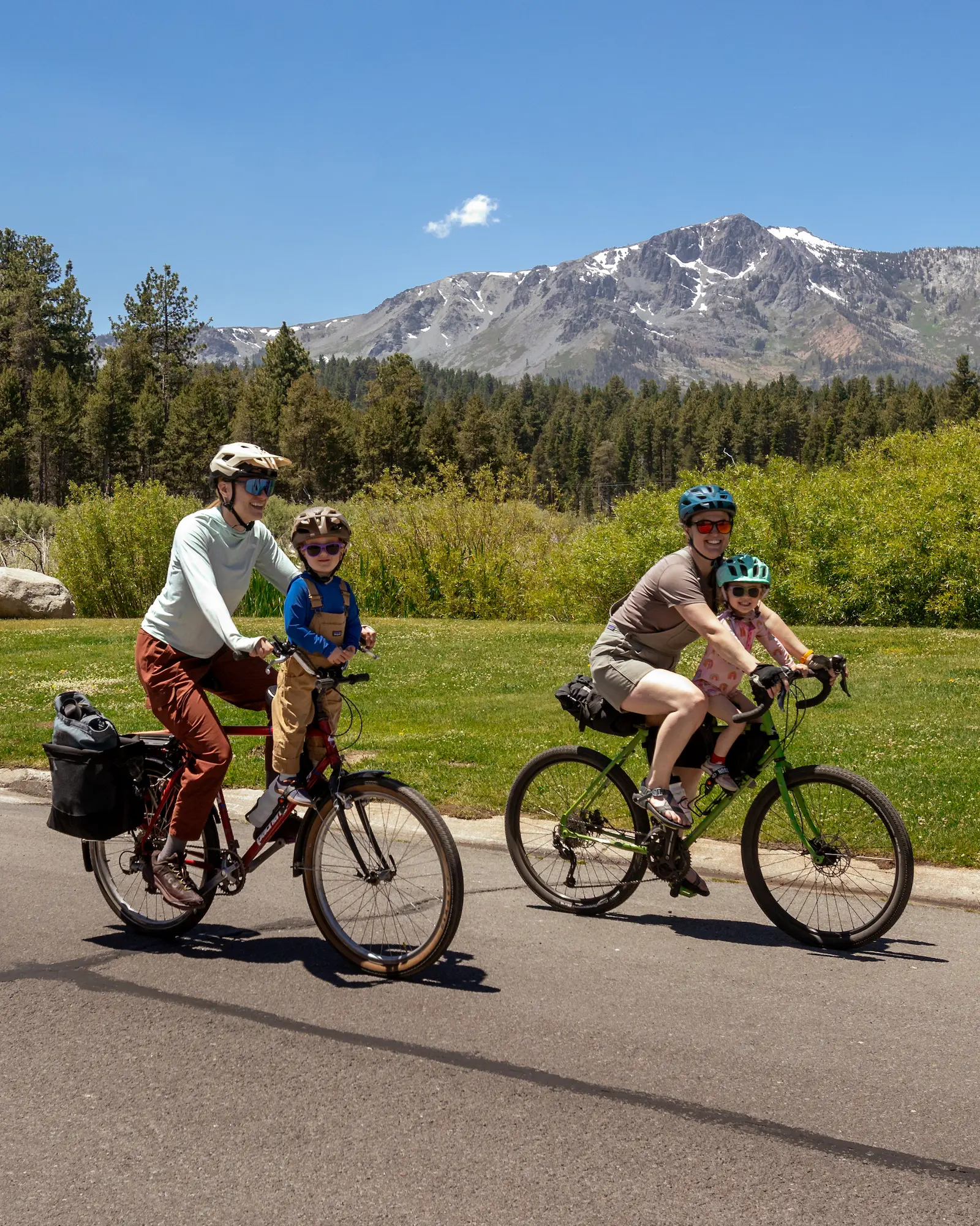 Two adult cyclists with small children on their bikes riding on a street with a large mountain in the background.