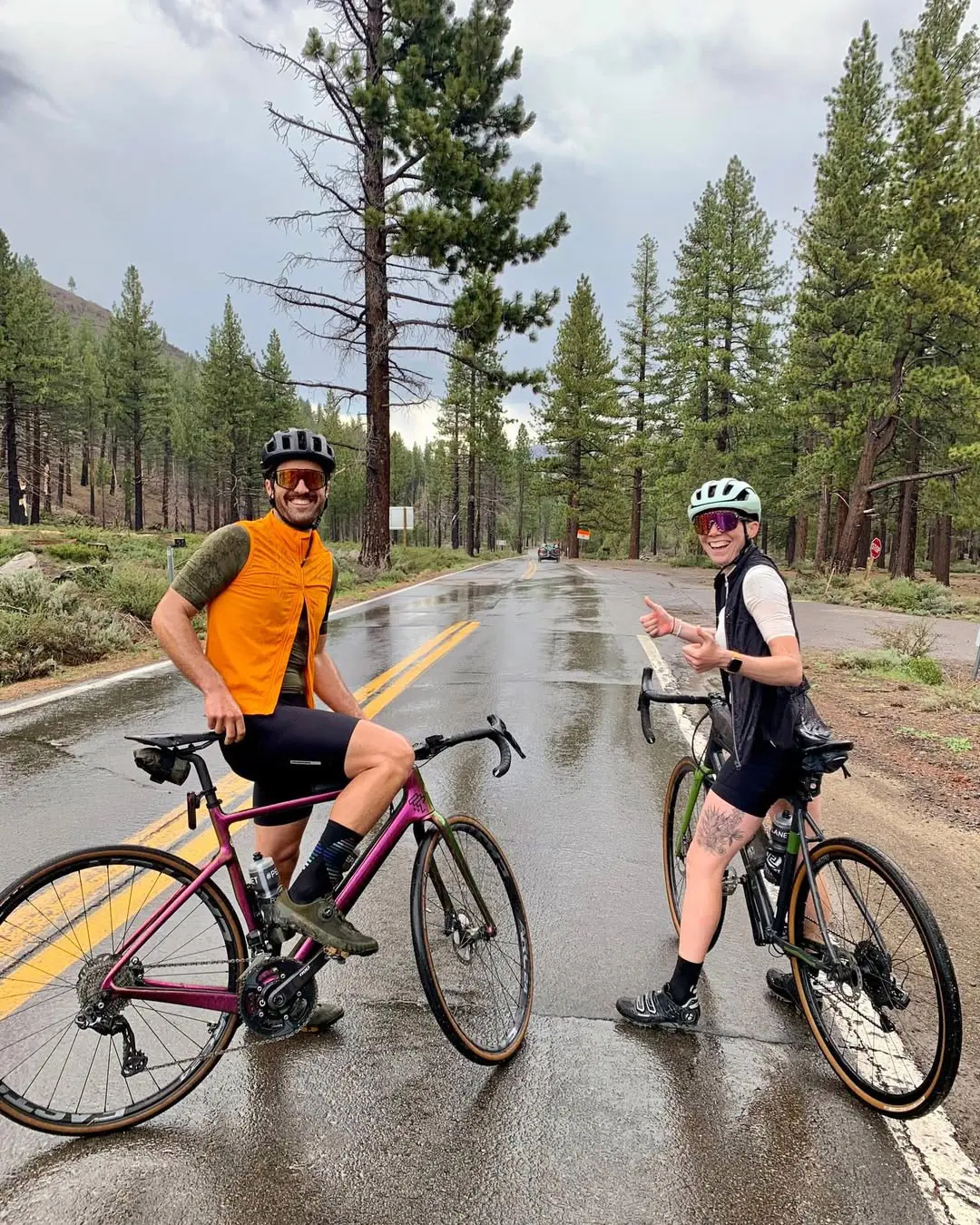 Two cyclists posing with their bikes on a rainy day surrounded by trees and mountains.